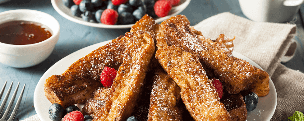 French Toast Sticks with Berries and  Topped with Powdered Sugar on a Plate with a Side of Syrup in a Bowl