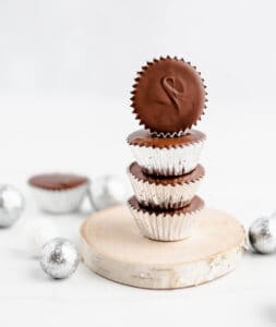 Three Peanut Butter Cups in Silver Liners with a Fourth Peanut Butter Cup Tilted on One Side All on a Wood Coaster with Silver Ball Decoration on the Table
