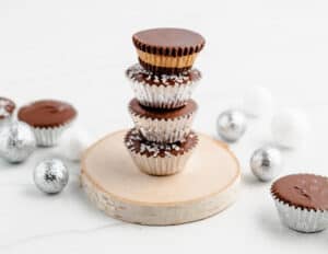 Four Stacked Peanut Butter Cups with Three in Silver Mini Cupcake Liners and the Fourth on Top Showing the Middle Layer of Peanut Butter Filling all on a wood coaster with silver ball decorations on the table