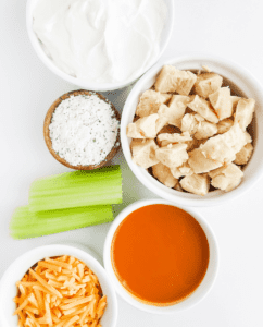Buffalo Chicken Dip Ingredients Including a Bowl of Sour Cream, Ranch Dressing or Dip Seasoning Mix, Cubed Cooked Chicken, Celery Sticks, Buffalo Sauce, and Shredded Cheese