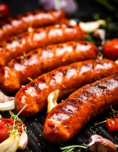 Grilled Bratwursts on a Dark Grilling Surface with Fresh Garlic, Herbs, and Veggies
