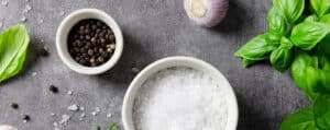 White Bowls of Salt, Pepper, and Garlic with Fresh Herbs on a Dark Grey Surface