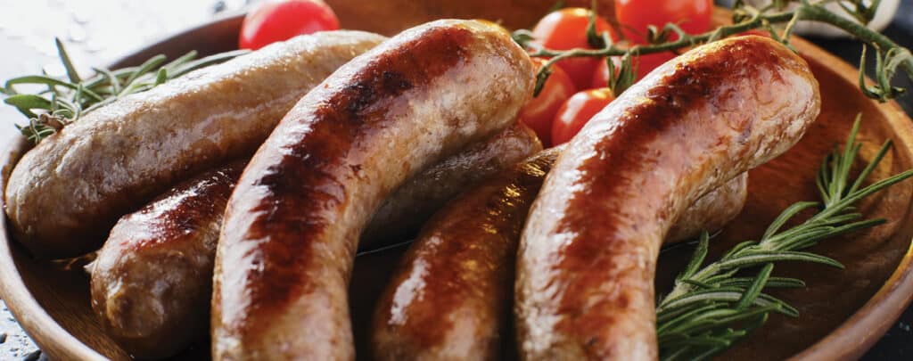 Pile of Cooked Bratwursts with Fresh Herbs and Tomatoes in a Wooden Bowl