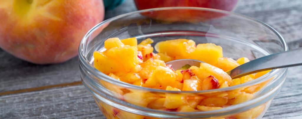 Fresh Diced Peached in a Clear Bowl with a Serving Spoon and Fresh Peaches on the Side on Wooden Surface