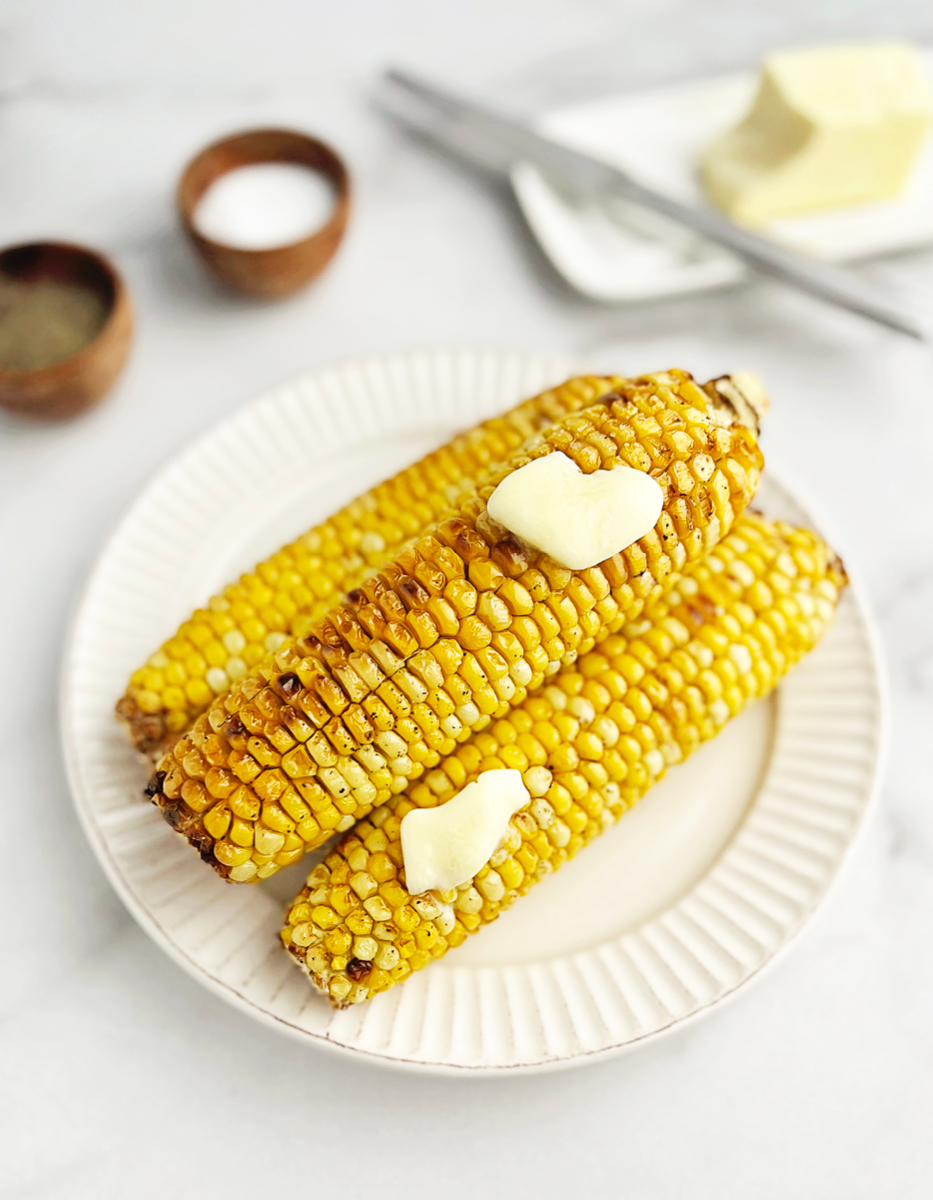 Air Fried Corn on the Cob on a Plate with Butter, Surrounded by Pepper, Salt, and Butter on a Plate with a Knife