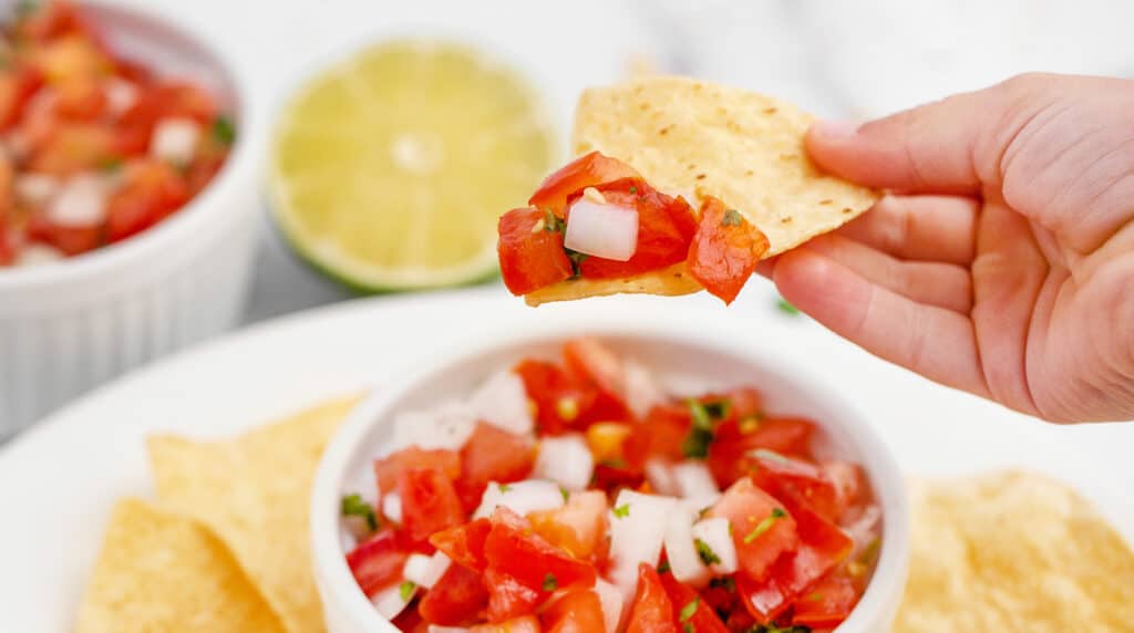 Scooping a Chip with Pico de Gallo with Bowl of Pico, Tortilla Chips, and Lime in the Background