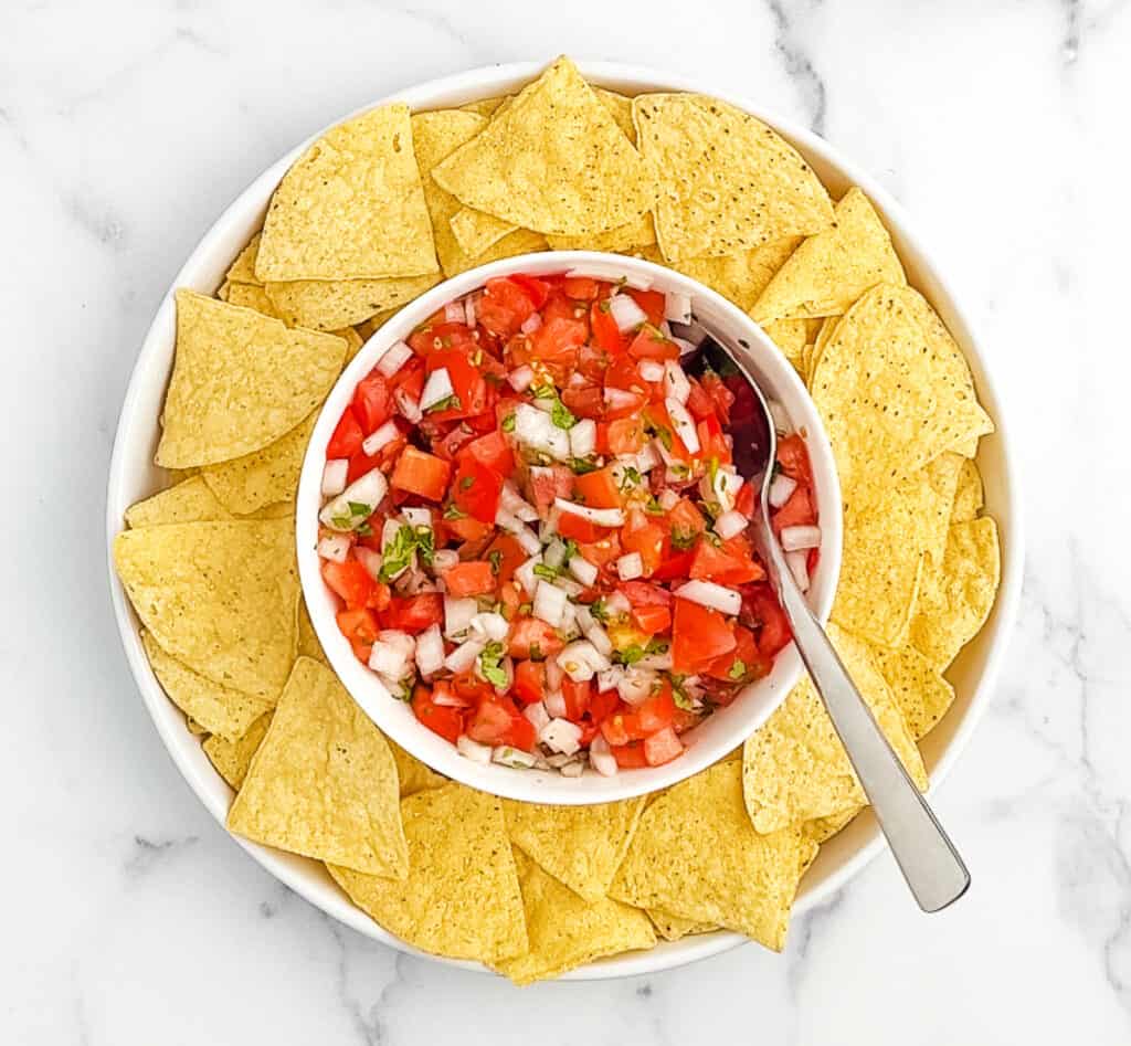 Fresh Pico de Gallo in Bowl with Circle of Tortilla Chips Around It on White Marble Surface