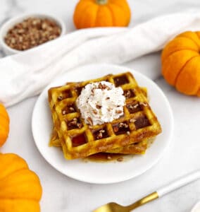 Pumpkin Waffles on a Plate with Whipped Cream, Syrup, and Spices with Mini Pumpkins on a White Surface