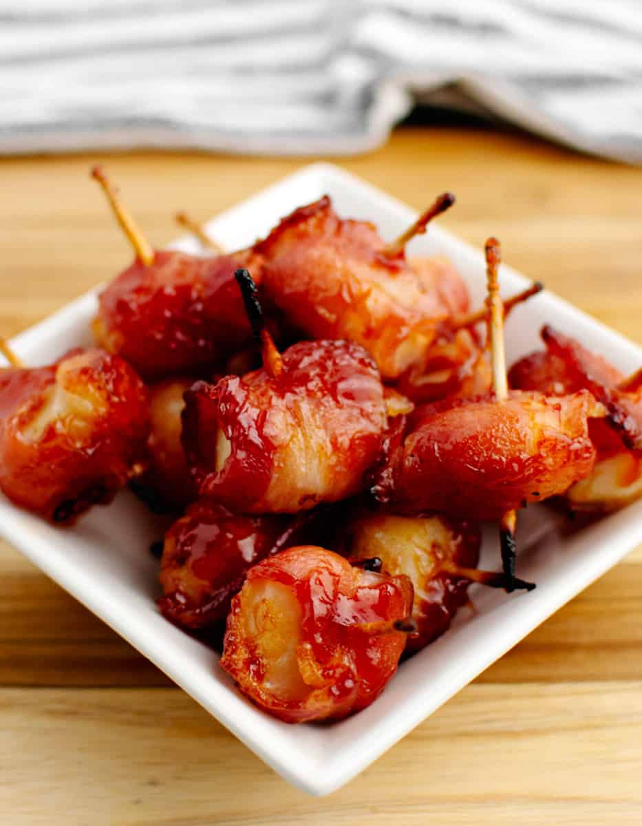 Bacon Wrapped Water Chestnuts Secured with Toothpicks in Square Bowl on Wooden Surface with Kitchen Towel in the Background