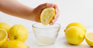 Squeezing Lemon Juice into Small Glass Bowl with Whole Lemons White Surface