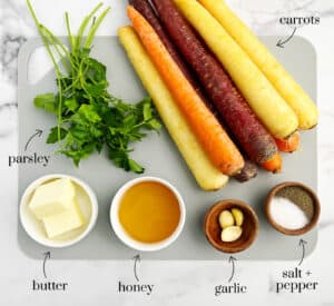 Rainbow Carrots, Parsley, Butter, Honey, Garlic, and Salt and Pepper on Grey Cutting Board on White Marble