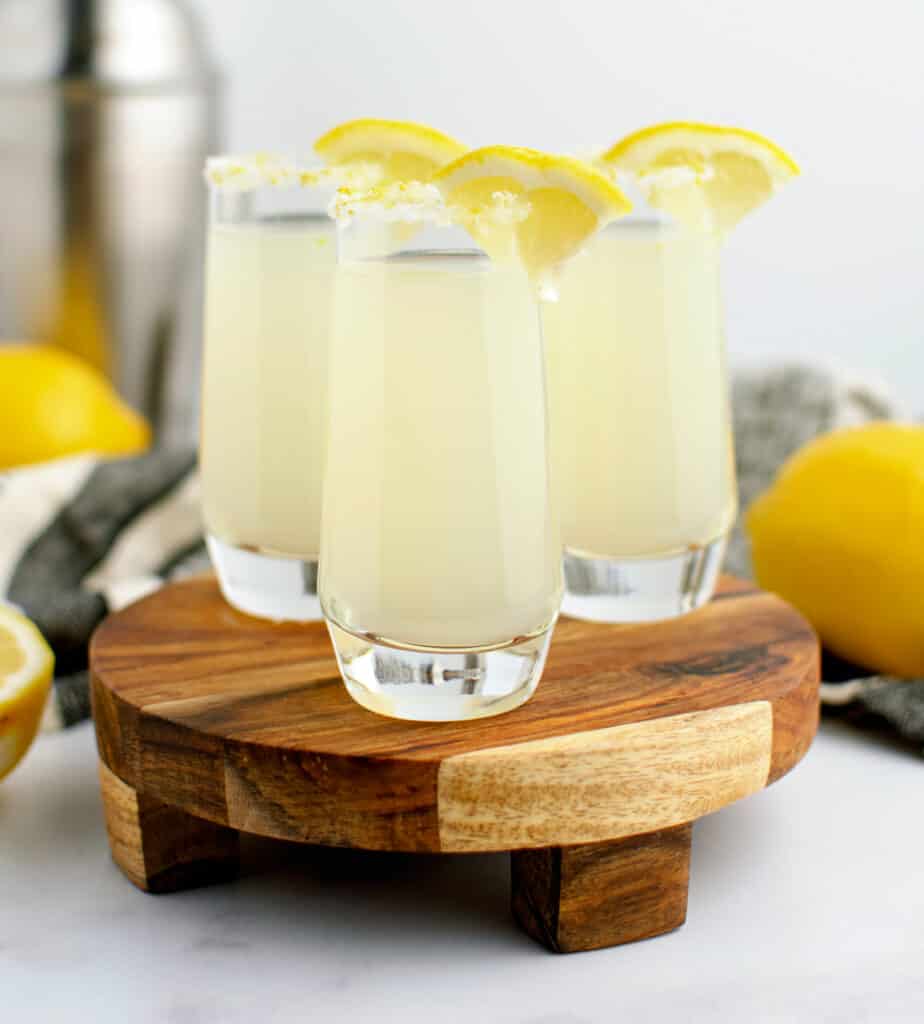 Lemon Drop in Shot Glasses with Wedge Garnish on Wooden Surface with Lemons and Cocktail Shaker in Background