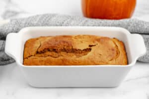 Side View of Pumpkin Loaf in White Pan with Grey Kitchen Towel and Pumpkin Behind