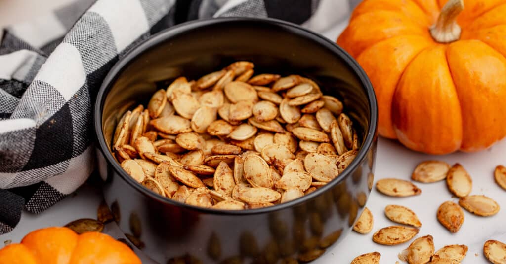 Air Fried Pumpkin Seeds in Black Bowl with Pumpkin and Kitchen Towel in the Background