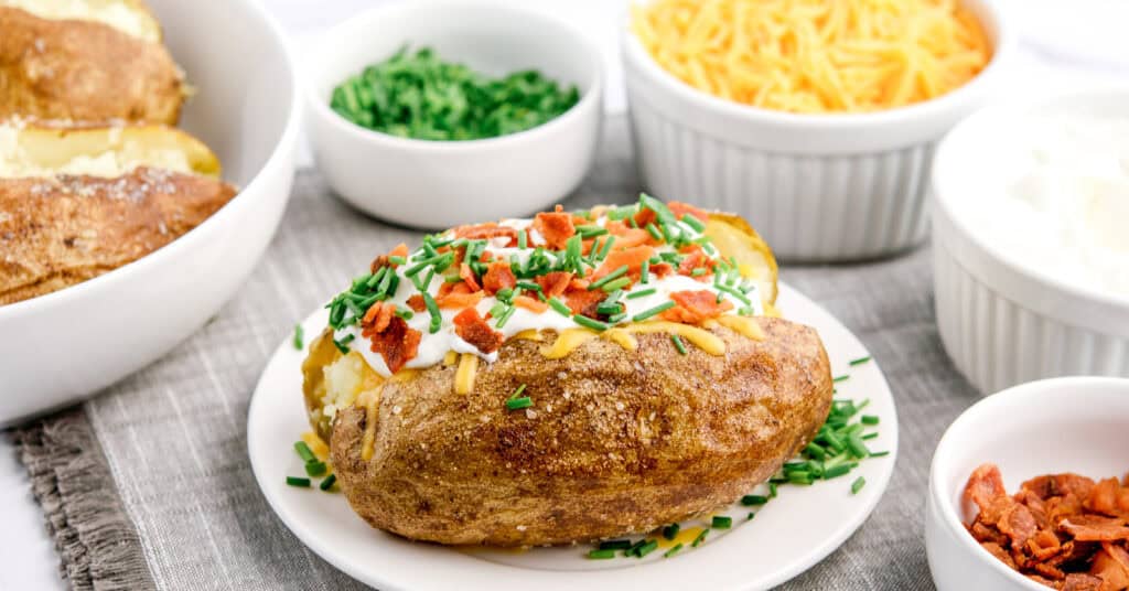 Baked Potato with Sour Cream, Cheese, Green Onions, and Bacon with Bowls of Toppings in the Back