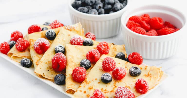 Crepes on a Plate with Blueberries and Raspberries Dusted with Powdered Sugar and Bowls of Berries Behind